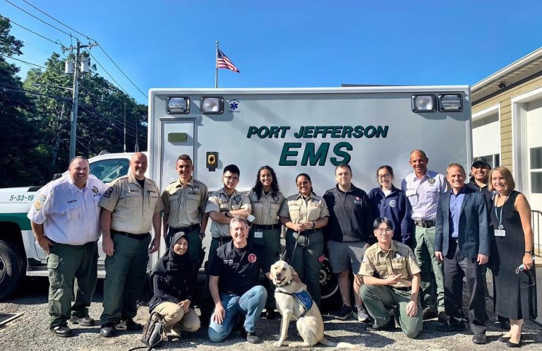 A group of people, including EMS staff and volunteers, pose in front of a Port Jefferson EMS vehicle. A service dog sits at the center. The American flag is visible in the background.