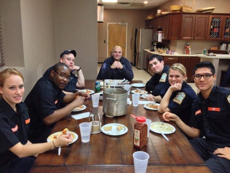 A group of six people in uniform sit around a wooden table, enjoying a meal. The table is set with plates, cups, and a large pot. The room is a kitchen or dining area with dishes and appliances in the background.