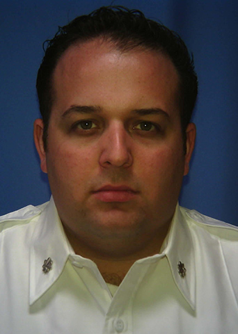 A person with short dark hair wearing a white shirt with decorative insignia on the collar poses against a plain blue background.