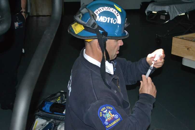 A paramedic in a dark uniform and safety helmet labeled "Physician MD 110" prepares a syringe. The patch on their sleeve reads "New York State Paramedic." They kneel on the floor in a dimly lit room with medical equipment nearby.