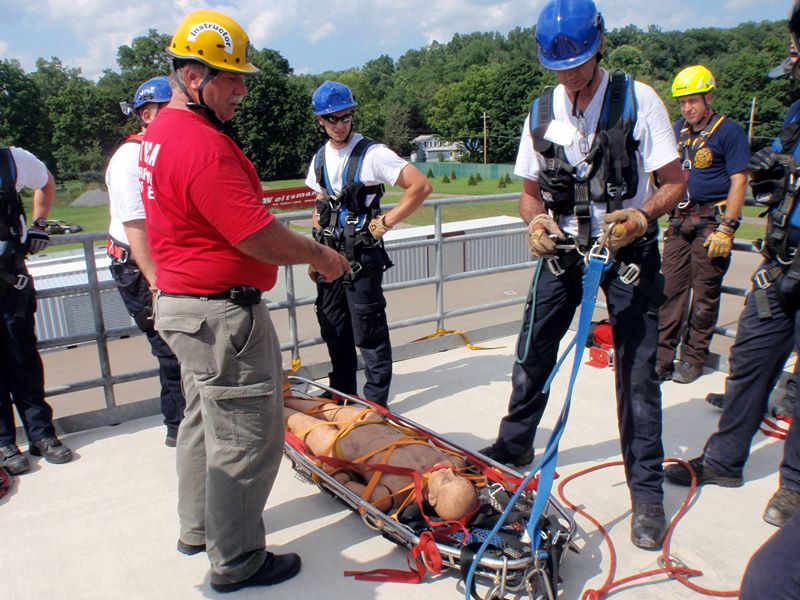 Rescue team conducts a high-rise training exercise. A person in protective gear is secured on a stretcher, while others in helmets and harnesses assist. The scene takes place on a rooftop with trees in the background.