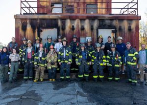 A group of firefighters wearing uniforms and helmets stand in front of a fire training structure. The team, consisting of both men and women, is posing for a group photo on asphalt with the building's metal exterior in the background.