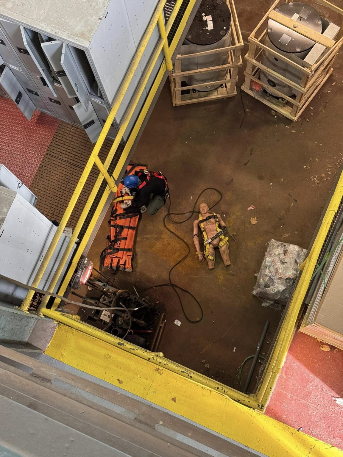 A worker in a blue helmet crouches next to safety equipment and orange cones on a rooftop. Below, a height safety dummy wearing a harness lies on the ground. The area is enclosed by yellow railings, with machinery and crates nearby.