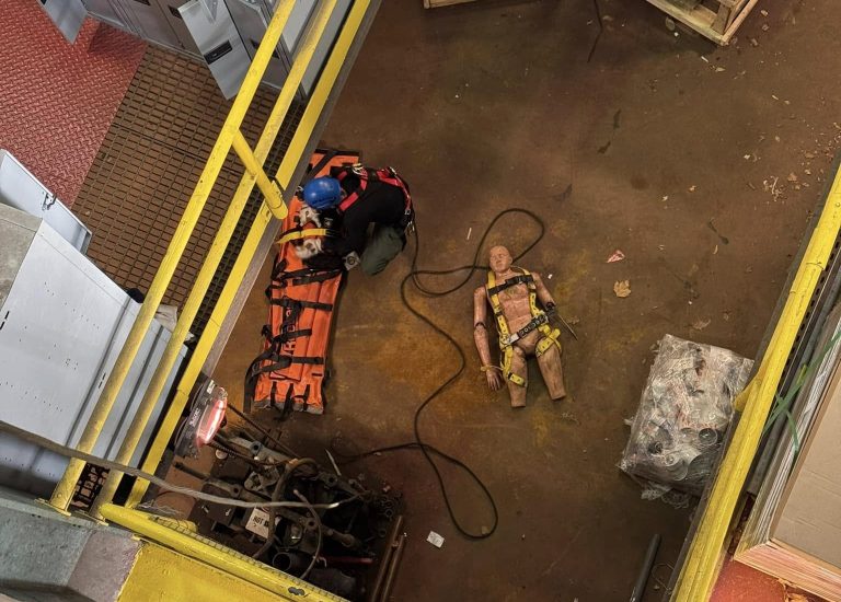 A worker in a blue helmet crouches next to safety equipment and orange cones on a rooftop. Below, a height safety dummy wearing a harness lies on the ground. The area is enclosed by yellow railings, with machinery and crates nearby.