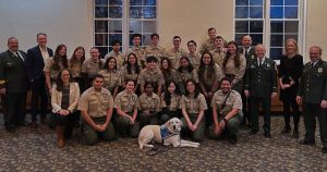 A large group of Boy Scouts in uniform, including young scouts, adult leaders, and a guide dog, pose indoors in front of tall windows. They are smiling and standing or kneeling. The setting includes a patterned carpet and cream-colored walls.