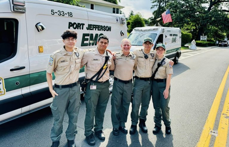 Five EMS personnel in uniform stand in front of an ambulance with "Port Jefferson EMS" on it. They pose smiling on a sunny day. An American flag is visible on the ambulance. Trees and a house are in the background.