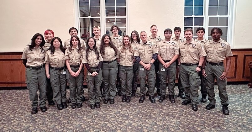 A group of people wearing beige shirts and green pants stand in two rows inside a room with carpeted flooring and large windows. They appear to be in a formal or official setting, standing closely together and smiling at the camera.