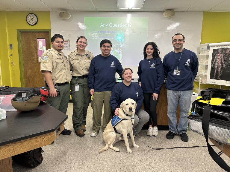Group of six people, including two in uniform, standing in a classroom. They are smiling and posing with a guide dog. A presentation screen behind them reads "Any Questions?". A model skeleton is visible on the right.