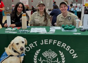 Three people in uniforms sit behind a table with "Port Jefferson" on the front. They are smiling and have promotional items on the table, including pens and green stress balls. A yellow Labrador wearing a blue vest sits in front of the table.