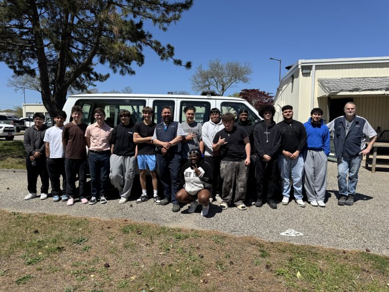 A group of sixteen people, mostly young men, stand together outdoors in front of a white van on a sunny day, posing and smiling for the photo. Trees and buildings are visible in the background.