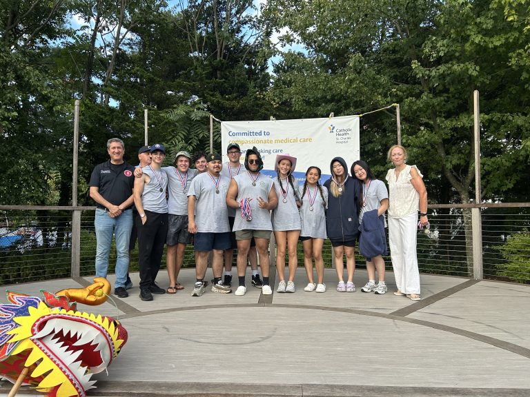 A group of ten people stand on a wooden stage outdoors, wearing medals and casual clothes. Behind them is a banner with healthcare messaging, and in the front left corner is a colorful dragon boat head. Trees fill the background.