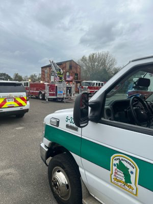 Emergency vehicles and personnel are gathered near a building with a ladder extended to its roof. Firefighters are on the scene under a cloudy sky, and a marked van is in the foreground with its door open.