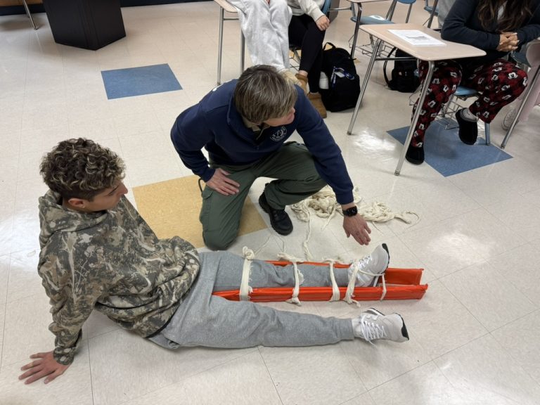 A person in a camouflage hoodie sits on the floor with their leg in a bright orange splint, secured with ropes. Another person kneels beside them, demonstrating the splint in a classroom setting.