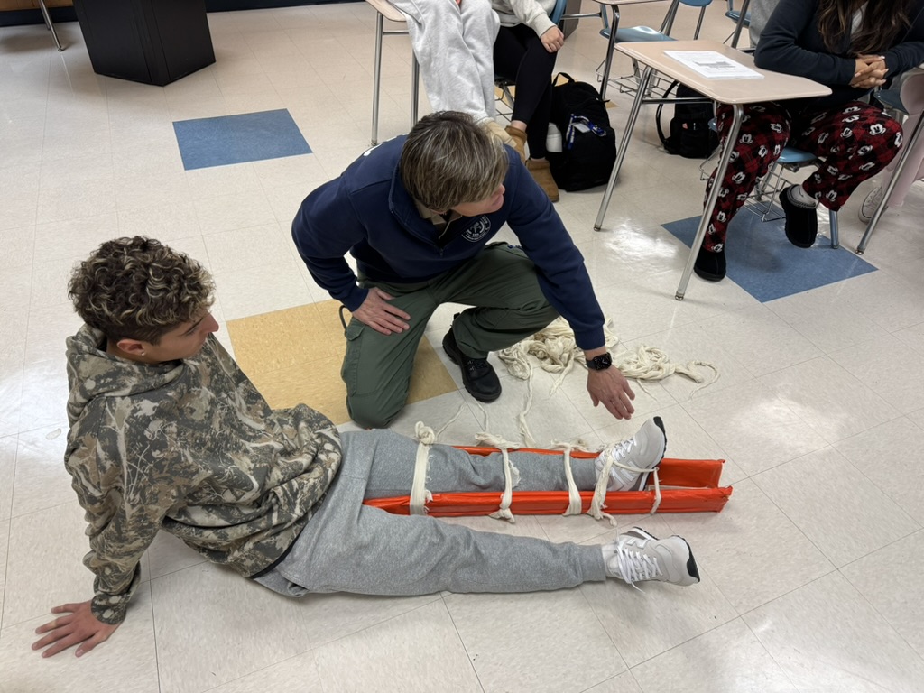 A person in a camouflage hoodie sits on the floor with their leg in a bright orange splint, secured with ropes. Another person kneels beside them, demonstrating the splint in a classroom setting.
