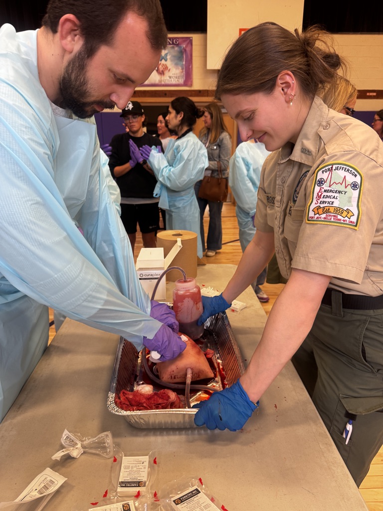 Two people wearing gloves and protective clothing handle a large animal organ in a metal tray on a table, surrounded by medical supplies, while others observe in the background inside a gymnasium.