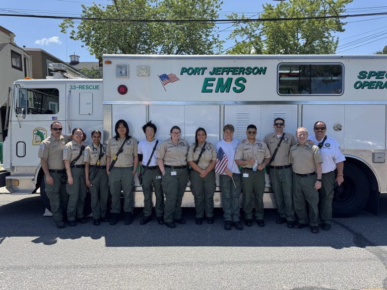 A group of twelve EMS personnel in uniform stand smiling in front of a Port Jefferson EMS vehicle on a sunny day. Trees and buildings are visible in the background.