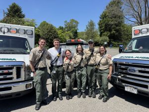 Six emergency medical personnel in uniform stand smiling together between two ambulances on a sunny day, with trees and blue sky in the background.