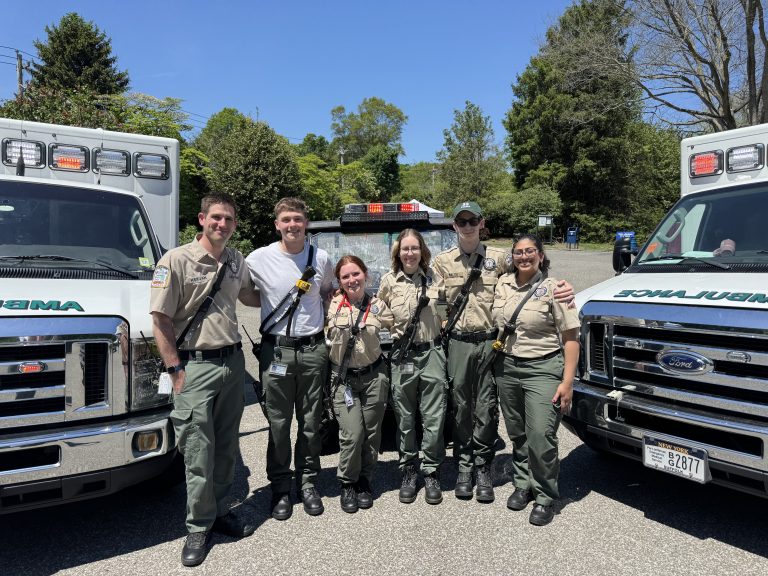 Six emergency medical personnel in uniform stand smiling together between two ambulances on a sunny day, with trees and blue sky in the background.